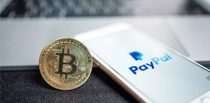 Bitcoin next to smartphone displaying PayPal logo with modern laptop in the background