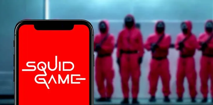 a phone with Squid Game on its screen and Squid Game guards standing behind it