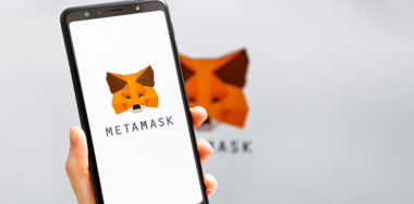Hand holding mobile with MetaMask app running at smartphone screen with MetaMask logo at background.