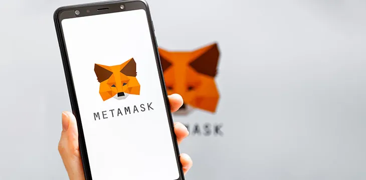 Hand holding mobile with MetaMask app running at smartphone screen with MetaMask logo at background.