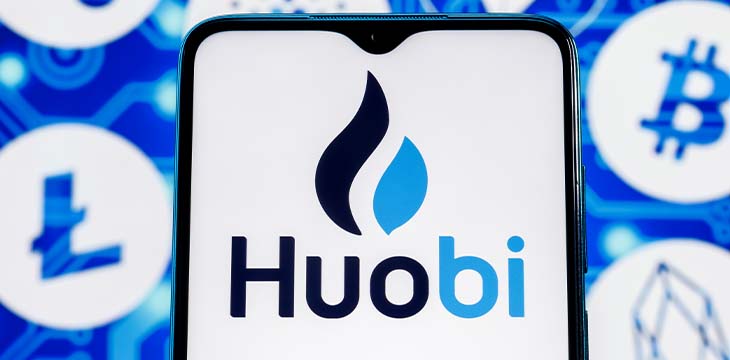 Huobi logo on smartphone screen against the background of the main cryptocurrencies.