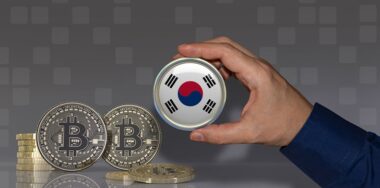 A hand holding a badge with the South Korean flag in front of some bitcoins. Crypto currency concept