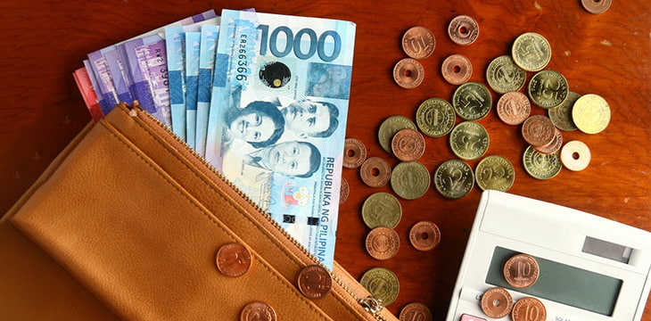 Philippine peso bills and coins in a leather wallet with a calculator on a wooden table