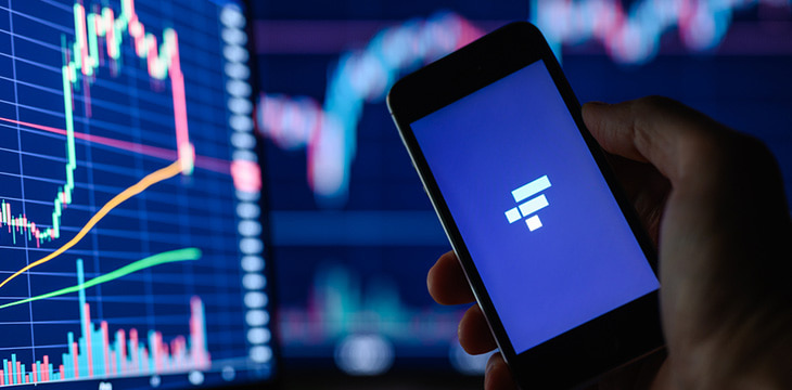 FTX cryptocurrency trading application on a smartphone in hand against the background of monitors with financial charts