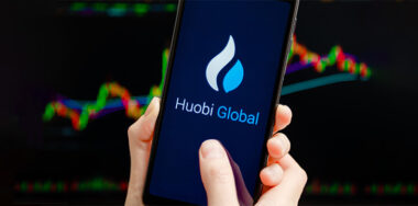 Hands holding mobile with Huobi Global app running at smartphone screen, trading candlestick chart at background. Huobi - cryptocurrency exchange, trading platform
