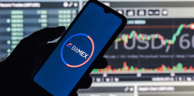 A smartphone with the BitMEX logo in hand on the background of the live trading webpage