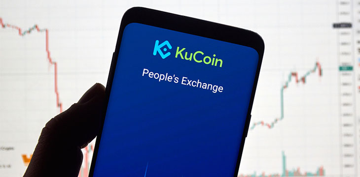 Kucoin, cryptocurrency exchange logo and application on Android Samsung Galaxy s9 Plus screen in a hand over a laptop display with bitcoin chart on it