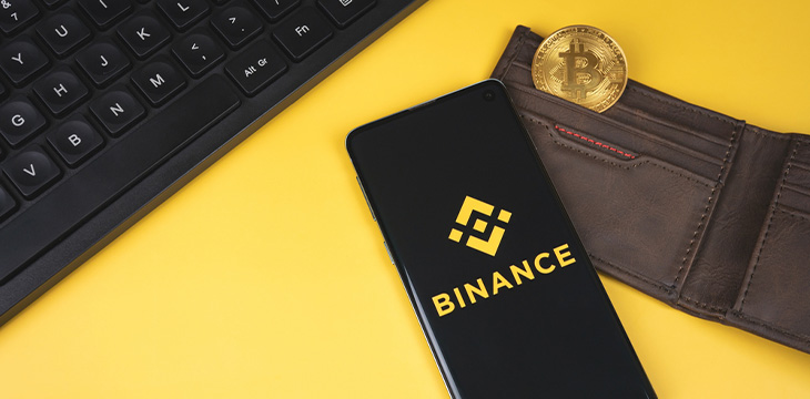 SWANSEA, UK - FEBRUARY 23, 2021: Binance mobile app logo on a smartphone, wallet with gold Bitcoin and keyboard on yellow background. Popular cryptocurrency exchange