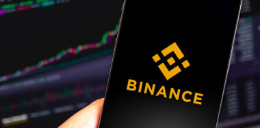 Binance logo mobile app on screen iPhone being held and MacBook screen in the background