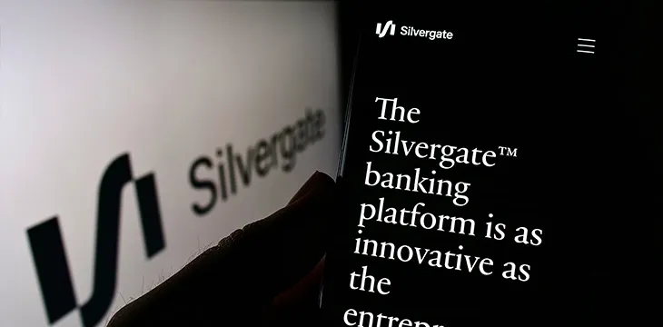 Hand holding the smartphone in front of the PC screen with the symbol of Silvergate