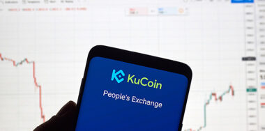 Kucoin exchange logo and application on Android Samsung Galaxy s9 Plus screen in a hand over a laptop display with chart