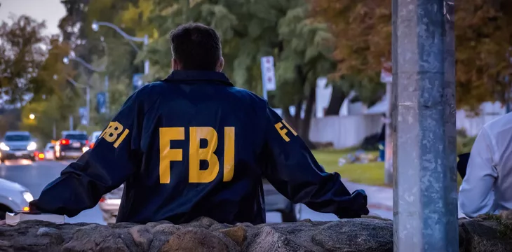 Male FBI agent wearing dark blue coat with FBI logo looking down the street with cars in the dusk seen from behind