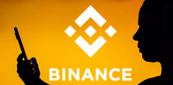 Binance logo on a yellow background with silhouette of a woman holding a smartphone