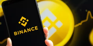 Binance mobile app running at smartphone screen with Binance logo at background.