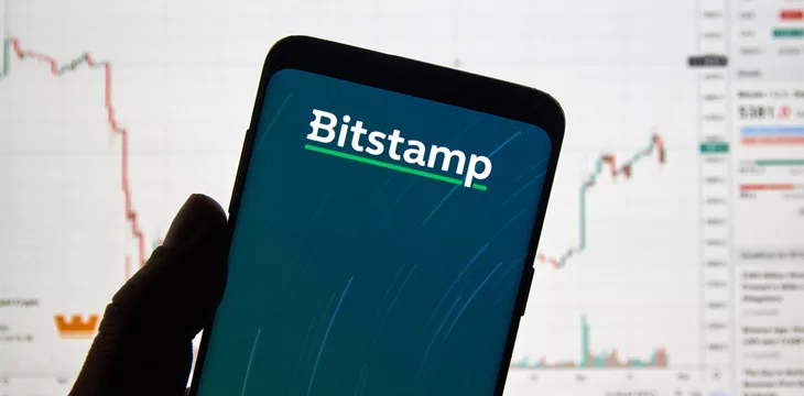 Bitstamp cryptocurrency exchange logo and application on Android Samsung Galaxy s9 Plus screen in a hand over a laptop display with bitcoin chart on it.