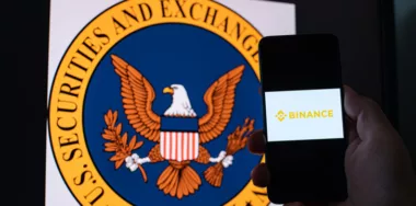 Man holds a smartphone with crypto exchange Binance on screen against the background of monitor with the SEC logo