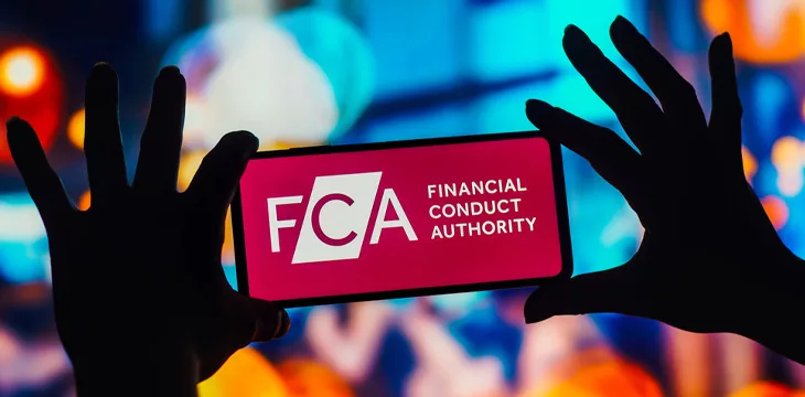 Financial Conduct Authority (FCA) logo is seen displayed on a smartphone being held by silhouette hands