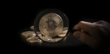 Unrecognizable person holding magnifying glass and looking through it on new virtual money golden bitcoin placed on a black background