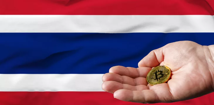 a hand holding a gold bitcoin in front of the flag of Thailand