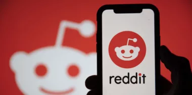 Reddit logo displayed on a smartphone device held by a silhoutted hand with Reddit logo on the background