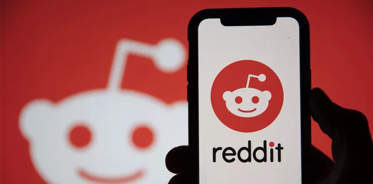 Reddit logo displayed on a smartphone device held by a silhoutted hand with Reddit logo on the background