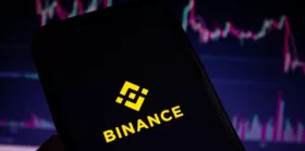 Binance mobile app logo displayed on the smartphone screen with blurry stock charts in the background.