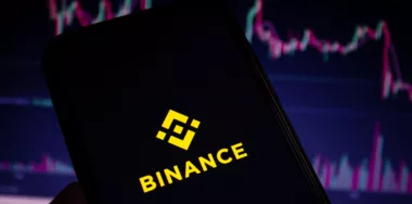 Binance mobile app logo displayed on the smartphone screen with blurry stock charts in the background.