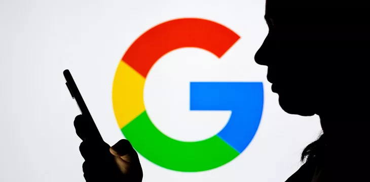 Google logo seen in the background of a silhouette woman holding a mobile phone