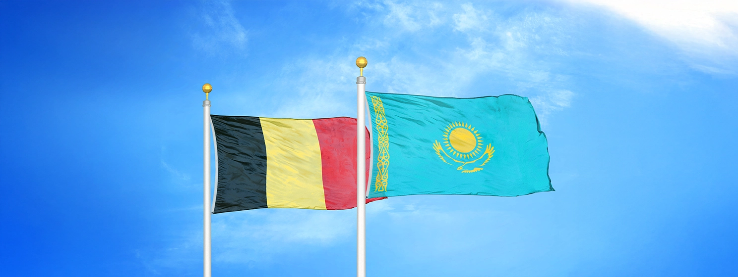Belgium and Kazakhstan two flags on flagpoles and blue cloudy sky background.