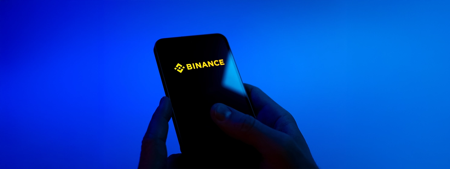 Hands on a blue background are holding a mobile phone displaying the Binance logo. Binance is a cryptocurrency exchange for trading digital assets.